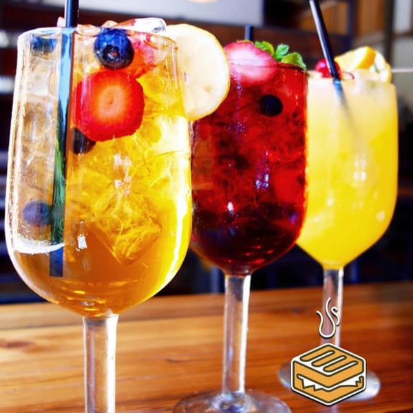 Three colorful fruit-garnished drinks in stemmed glasses on a wooden table at Dallas Grilled Cheese Co.
