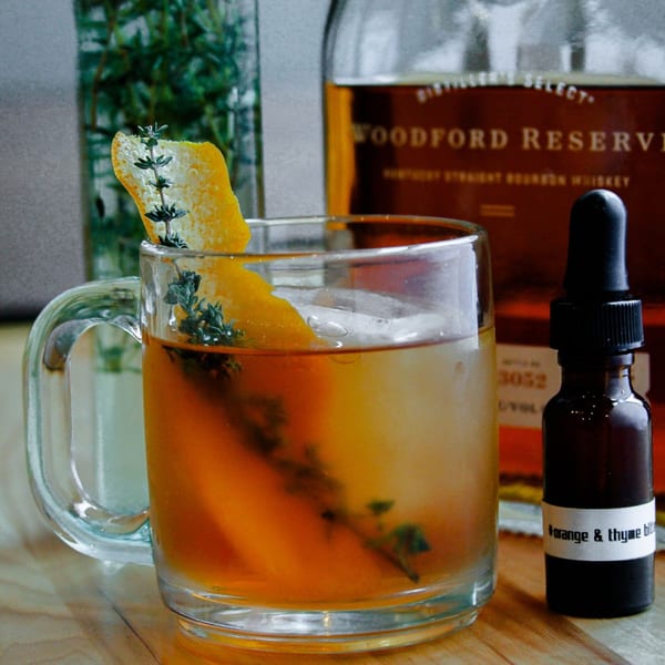 A whiskey cocktail with orange peel and thyme, next to a bottle of Woodford Reserve.