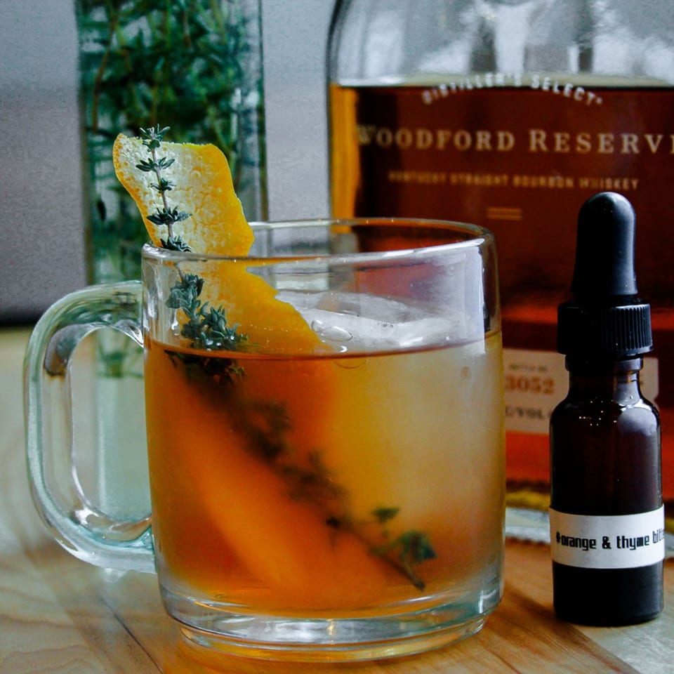 A whiskey cocktail with orange peel and thyme, next to a bottle of Woodford Reserve.